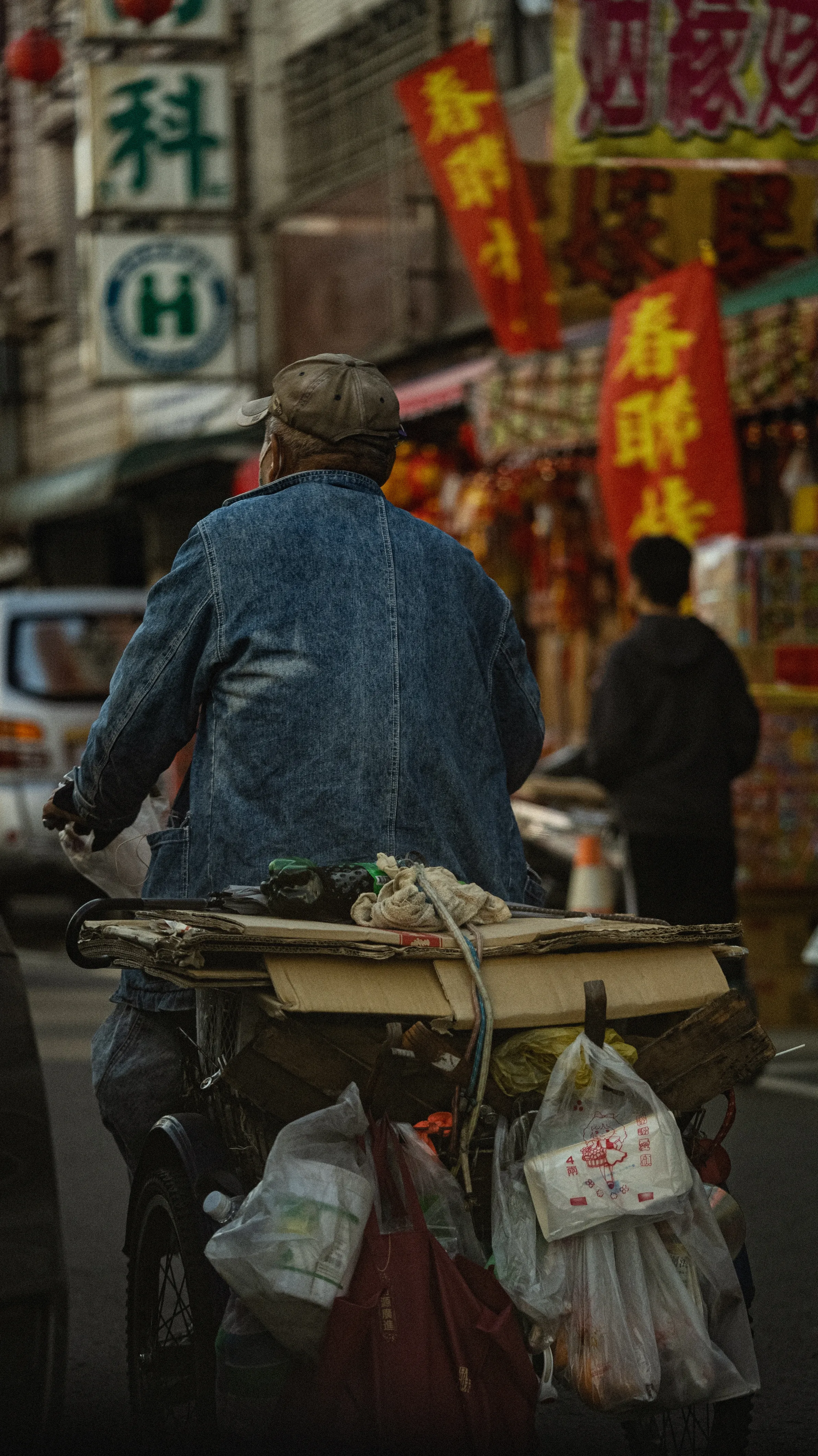 灰髮男子推著掛滿塑膠袋與雜物的載貨車,前方街道兩側布滿醒目招牌
