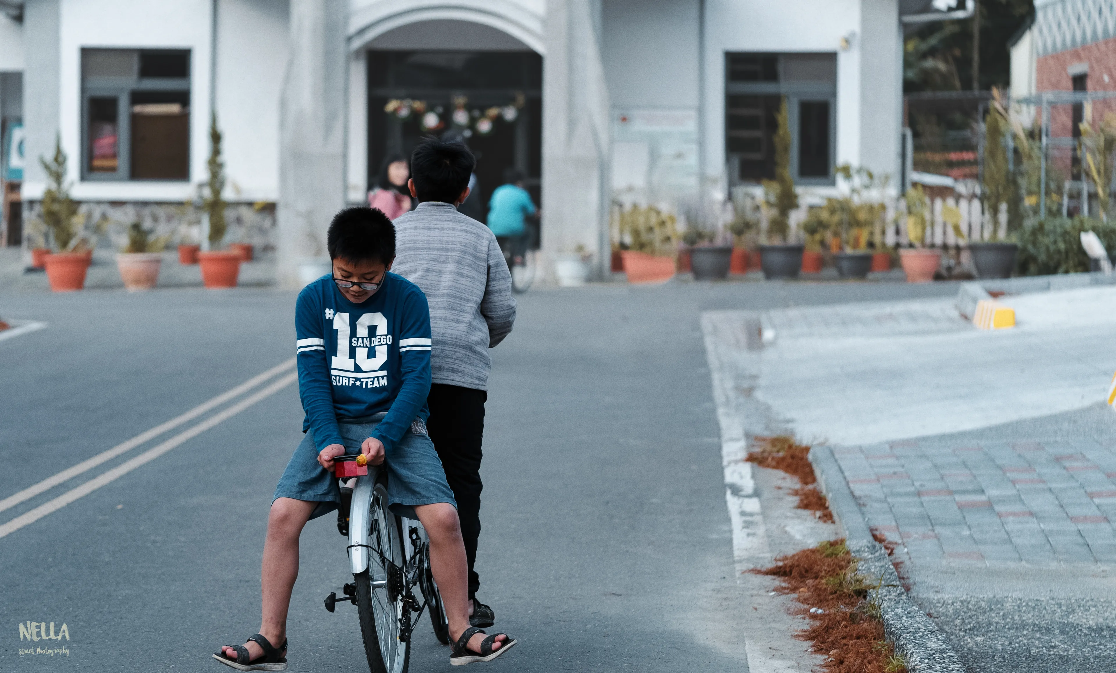 兩名孩童從後方騎腳踏車沿街道前進,前方是白色建築與施工錐