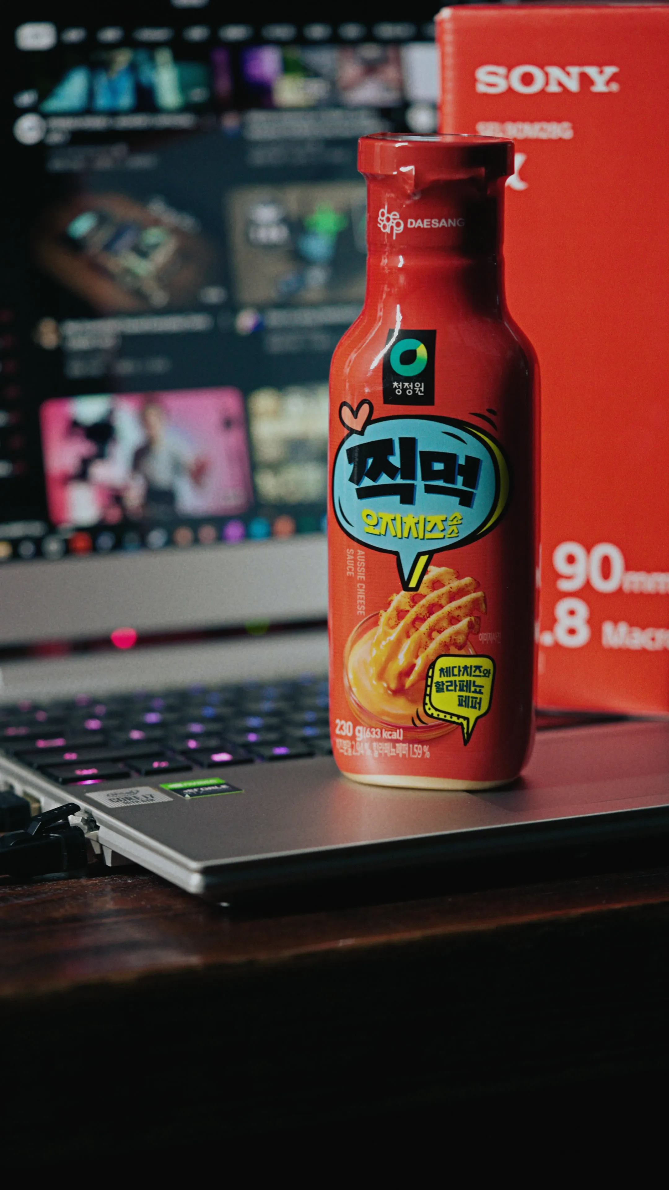 紅色瓶裝食品立在筆電鍵盤上,背景螢幕顯示影像編輯畫面並有紅色 Sony 包裝