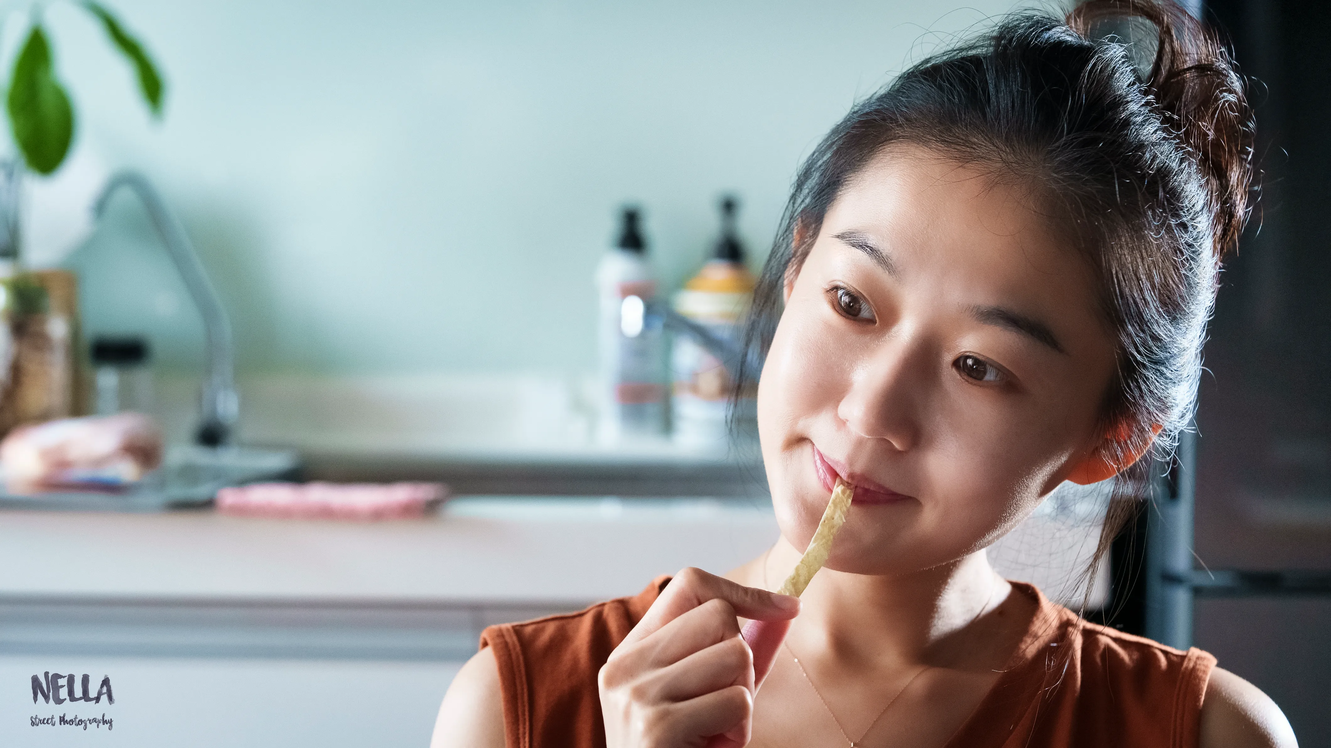 綁起頭髮的女子坐在廚房前,手持細長食物靠近嘴邊並望向側方